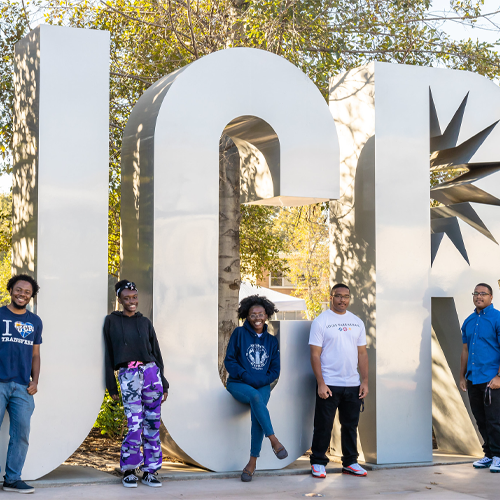 Group of UCR students stand right in front of the UCR campus sign on a sunny day.