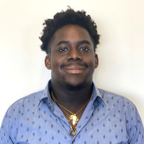 UCR student, Ayo Adeniran stands against a light background and smiles while wearing gold chain necklaces and light blue shirt. 