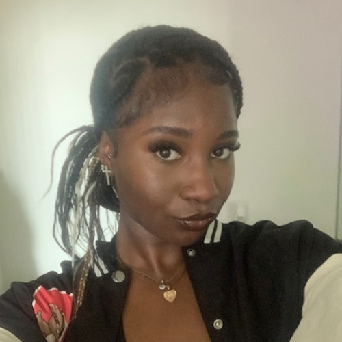 UCR student, Queen Udeh, stares at the camera while wearing a black and white jacket, cross earrings and a gold heart-shaped pendant necklace